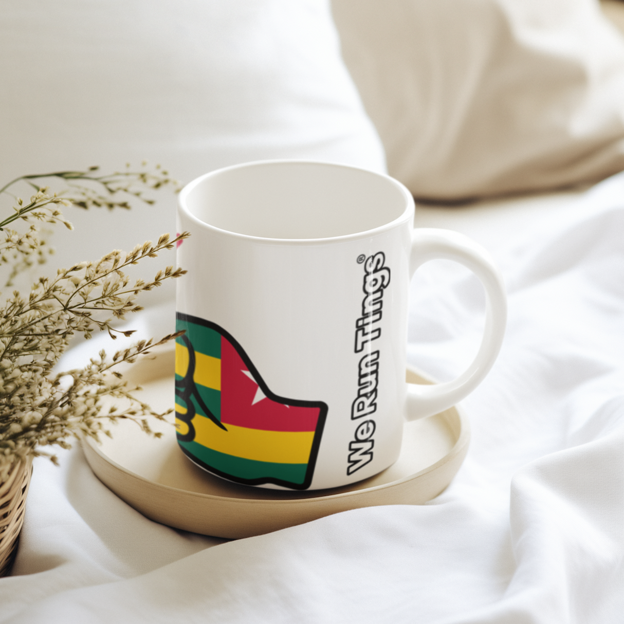 White ceramic “We Run Tings” mug featuring a raised fist in Togo flag colors, placed on a wooden coaster on soft white bedding, styled with delicate dried flowers for a calm, cozy lifestyle setting.