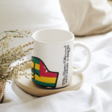 White ceramic “We Run Tings” mug featuring a raised fist in Togo flag colors, placed on a wooden coaster on soft white bedding, styled with delicate dried flowers for a calm, cozy lifestyle setting.