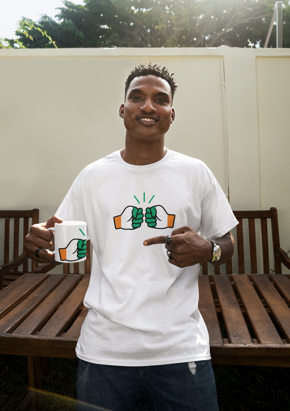 Man wearing a white Cote D'Ivoire t-shirt with a graphic design, holding a We Run Tings mug outdoors.