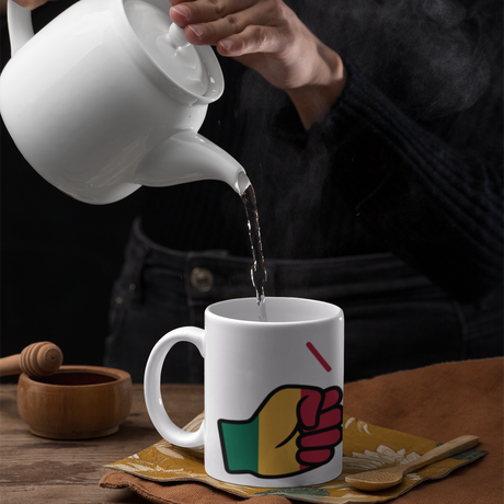 We Run Tings Mali white ceramic tea, coffee, mug, cup and white teapot pouring tea into the branded mug with a colorful design on a wooden table.