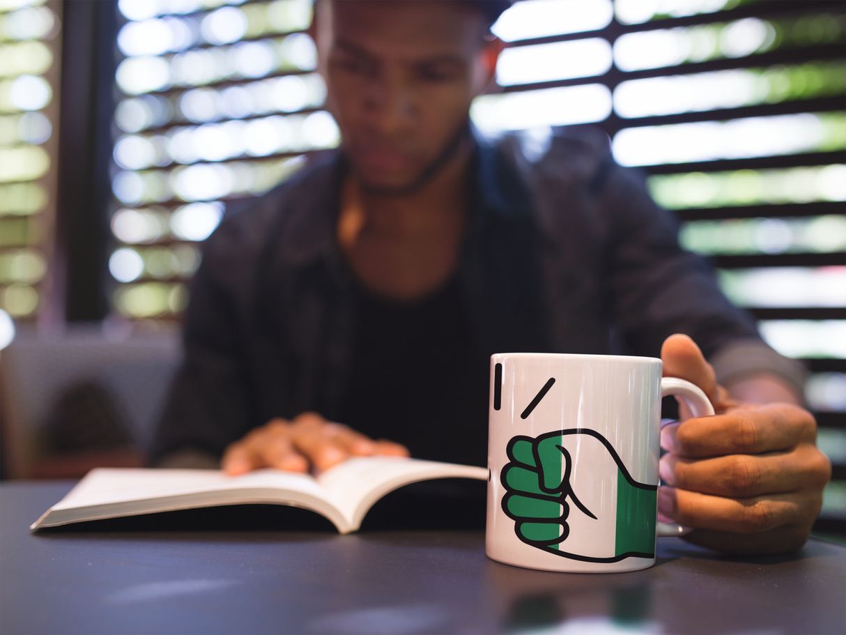Man holding a We Run Tings Nigeria white, ceramic, mug, cup with a fist symbol while reading a book in a blurred indoor setting