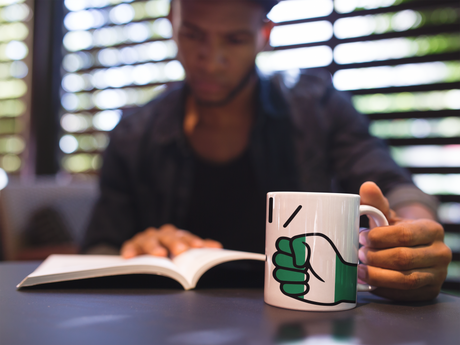 Man holding a We Run Tings Nigeria white, ceramic, mug, cup with a fist symbol while reading a book in a blurred indoor setting