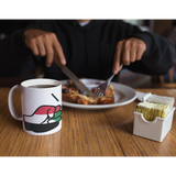 Person eating breakfast with a White We Run Tings, Sudan Mug with a graphic of a fist on a wooden table.