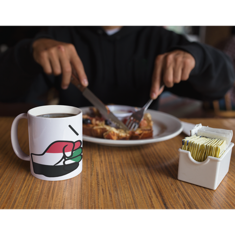 Person eating breakfast with a White We Run Tings, Sudan Mug with a graphic of a fist on a wooden table.