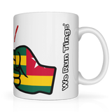 White ceramic “We Run Tings” mug featuring a raised fist wrapped in the Togo flag colors, with the brand name printed vertically on the side, symbolizing empowerment, unity, and cultural pride.