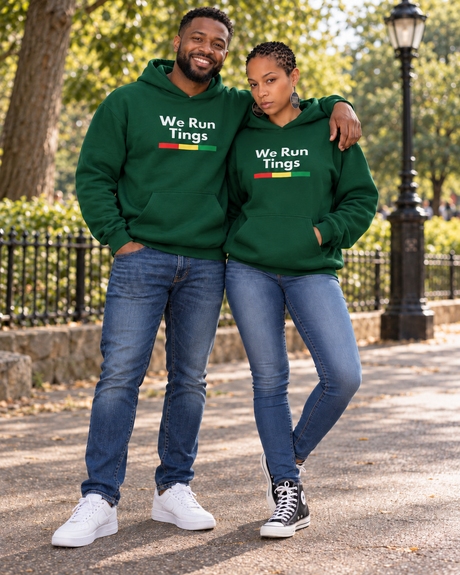 A man and a woman standing together outdoors on a park path, both wearing matching dark green hoodies with the white text 'We Run Tings' and a small red, yellow, and green stripe graphic. The man wears blue jeans and white sneakers, and the woman wears blue jeans and black-and-white sneakers.